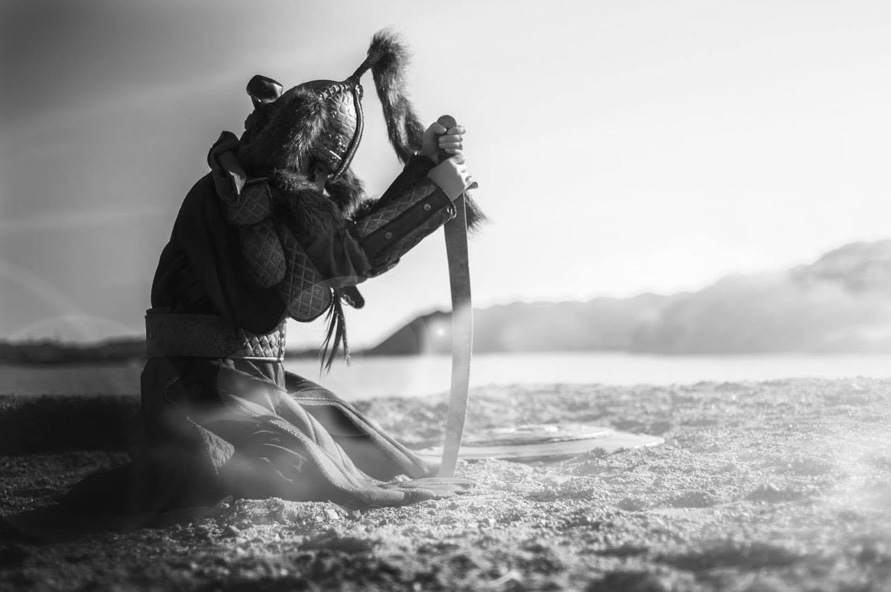 Black-and-white historical costume photograph: kneeling figure with a sword